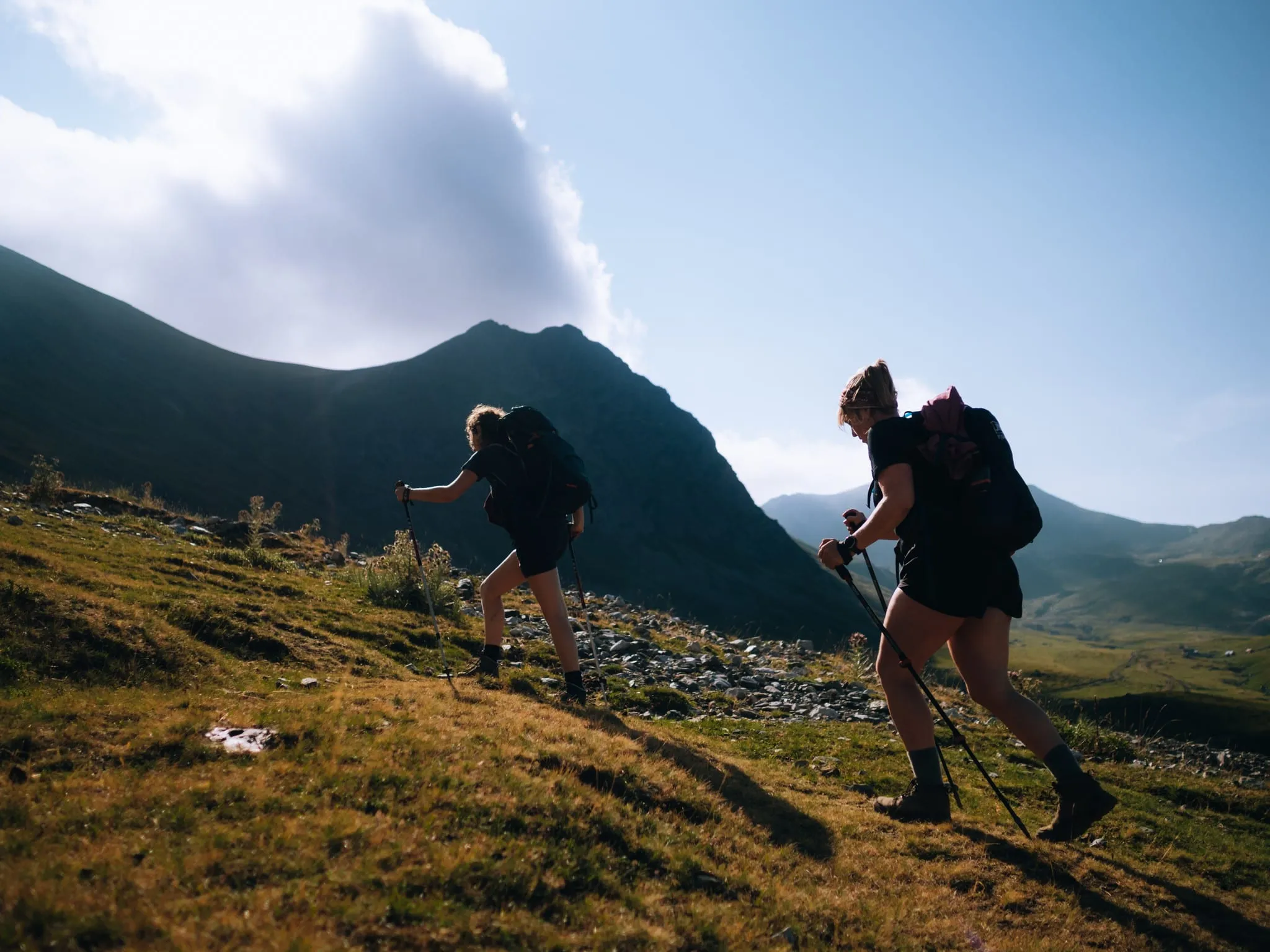 Deux randonneurs avec sacs à dos et bâtons montent une pente herbeuse ensoleillée, devant une chaîne de montagnes