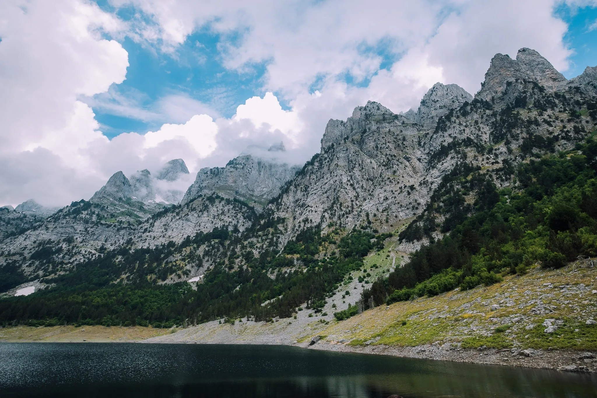 Un lac de montagne sombre sous des sommets calcaires déchiquetés enveloppés de nuages, dans le parc national de Theth